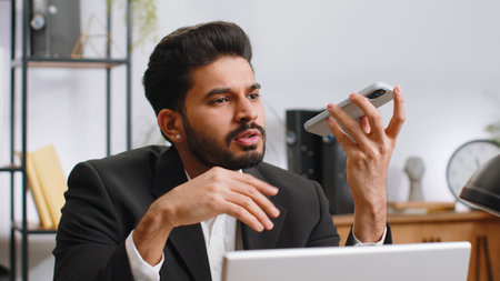 Indian businessman working, having mobile phone loudspeaker talk at home office desk with laptop. Bearded professional freelance man holding smartphone using messenger chat apps. Employment occupationの写真素材