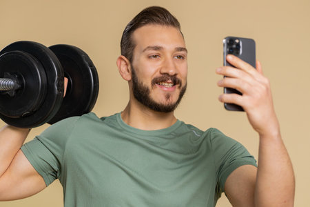 Athlete sportive man texting share messages content on smartphone social media applications online, sms message, browsing, reading news. Middle eastern sportsman guy with dumbbell on beige backgroundの写真素材