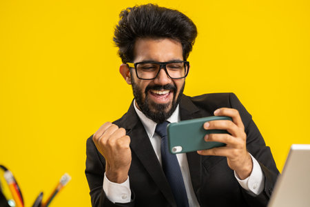 Indian businessman playing on smartphone exciting mobile application celebrate winning racing or shooter game at office desk workplace. Professional manager freelancer man enjoy good work of new appの写真素材