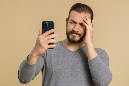 Lebanese man use mobile cell phone typing browsing, loses becoming surprised sudden lottery results, bad fortune, loss, fail. Handsome middle eastern guy isolated on beige studio background, indoorsの写真素材