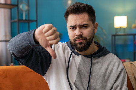 Dislike. Upset young man showing thumbs down sign gesture, expressing discontent, disapproval dissatisfied bad work at modern home apartment indoors. Displeased Caucasian guy in living room on sofaの写真素材