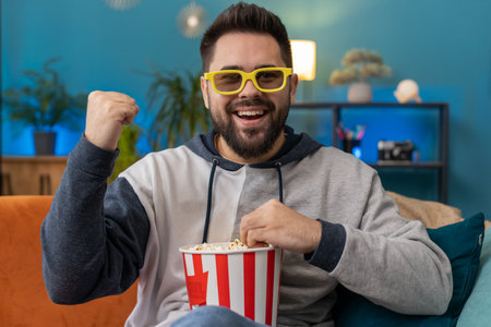 Excited young man in 3D glasses sits on sofa eating popcorn snacks and watching interesting TV serial sport game, film, online social media movie content at home apartment. Guy enjoying entertainmentの写真素材