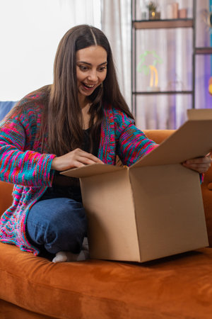 Happy young woman unpacking delivery parcel sitting at home room. Smiling satisfied Caucasian girl...