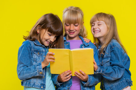 Teenage girls reading funny interesting fairytale story book, leisure hobby, knowledge wisdom, education, learning, study, wow. Little children sisters. Three cute siblings kids on yellow backgroundの写真素材