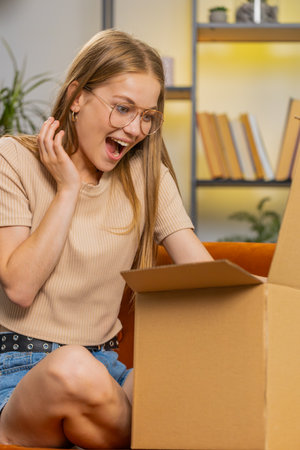 Happy woman unpacking delivery parcel sitting at home apartment on sofa. Satisfied girl shopper...