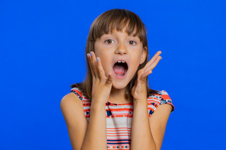 Oh my God, Wow. Young preteen child girl kid looking surprised at camera with big eyes, shocked by sudden victory game winning lottery goal achievemen good news. Children isolated on blue backgroundの写真素材