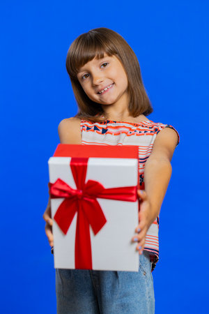 Lovely smiling preteen child girl kid presenting birthday gift box stretches out hands, offer wrapped present career bonus, celebrating party. Little cute children on studio blue background. Verticalの写真素材