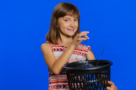 Young school girl taking off, throwing out glasses into bin after vision laser treatment therapy, looking smiling at camera. Healthy preteen Caucasian child kid isolated on studio blue backgroundの写真素材