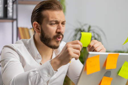 Tired exhausted young businessman working on laptop at office with many sticker task inscriptions. Freelancer man busy occupation feels sad boredom overworked multitasking. Panic attack, deadlineの写真素材