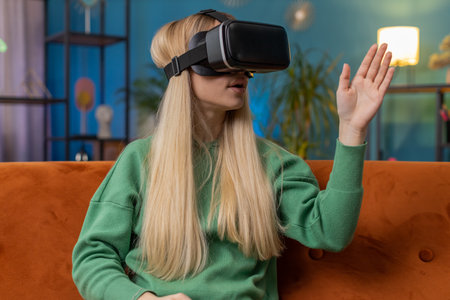 Excited woman using virtual reality futuristic technology VR app headset helmet to play simulation 3D 360 video game watching film movie. Child girl kid in goggles at modern home apartment on sofaの写真素材