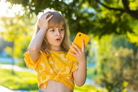 Preteen child kid play online game use smartphone typing browsing, loses becoming surprised by lottery results, bad news, fortune loss, fail. Blonde girl standing on city sunset park street, outdoorsの写真素材