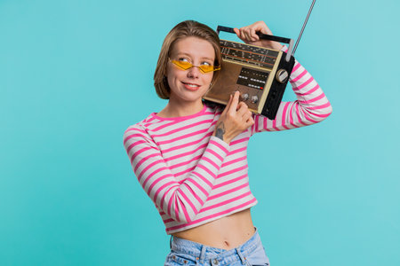 Woman in sunglasses using retro tape record player to listen music, disco dancing of favorite track, having fun, entertaining, fan of vintage technologies. Girl in crop top isolated on blue backgroundの写真素材