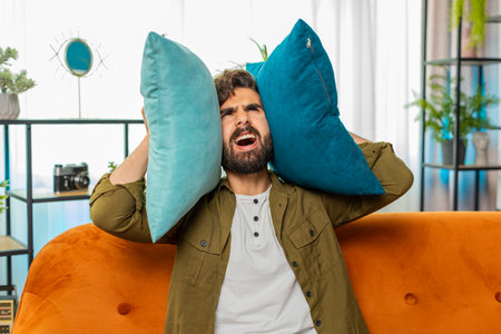 Repair work at neighbors. Irritated arabian man cover ears with pillows annoyed by noisy loud music neighbors suffer from headache wishes silence. Thin walls at home flat without sound insulationの写真素材