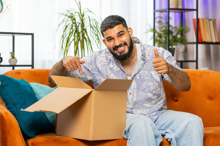 Happy Indian young man unpacking delivery parcel sits at home. Smiling satisfied bearded Arabian guy shopper online shop customer opening cardboard box receive purchase gift by fast postal shippingの写真素材