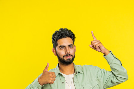 Indian bearded man showing thumbs up and pointing overhead, above head empty place, advertising area for commercial text copy space for goods promotion advertisement. Guy isolated on yellow backgroundの写真素材