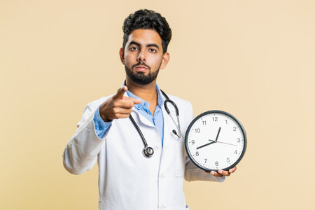 It is your time to heal. Indian young doctor cardiologist man showing time on wall office clock, ok, thumb up, approve, pointing finger at camera. Arabian apothecary pharmacy guy on beige backgroundの写真素材
