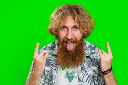 Overjoyed happy young caucasian man showing rock n roll gesture by hands, cool sign, shouting yeah with crazy expression dancing, rejoicing victory, win, success. Guy on green chroma key backgroundの写真素材