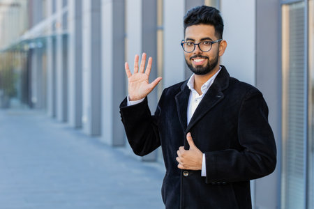 Hello. Indian businessman smiling friendly at camera, waving hands gesturing hi, greeting or goodbye welcoming with hospitable expression outdoors. Arabian Hindu freelancer guy in downtown city streetの写真素材