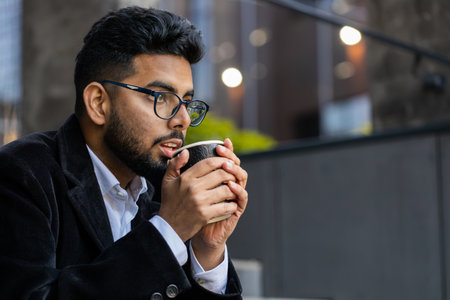 Happy bearded Indian businessman enjoying morning coffee hot drink and smiling outdoors. Relaxing, taking a break. Arabian Hindu freelancer guy drinking coffee to go in downtown city street. Lifestyleの写真素材