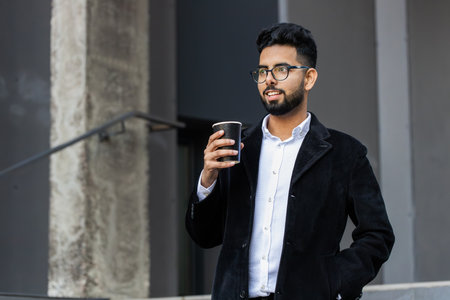 Happy bearded Indian businessman enjoying morning coffee hot drink and smiling outdoors. Relaxing, taking a break. Arabian Hindu freelancer guy drinking coffee to go in downtown city street. Lifestyleの写真素材