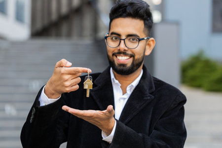Indian businessman real estate agent lifting hand showing keys of new home house apartment buying or renting property, mortgage loan, investment outdoors. Arabian Hindu freelancer guy in downtown cityの写真素材