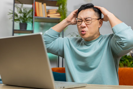 Displeased sad Asian man use laptop notebook typing browsing working, loses becoming surprised...