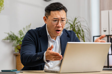 Irritated tired Asian business man while working on laptop, unexpected online website problem, computer virus data loss by hacking. Freelancer feeling mad about broken notebook at office workplaceの写真素材