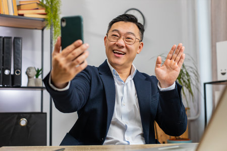 Happy excited Asian middle-aged businessman blogger influencer making phone conversation call with social media followers at office workplace working on laptop. Freelancer Chinese man in formal suitの写真素材