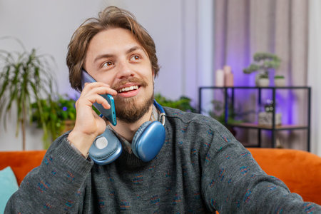 Phone call, good news, gossip. Happy surprised smiling man in pleasant conversation on smartphone, enjoying talking with friend indoors. Caucasian young guy sitting at sofa workplace home office roomの写真素材