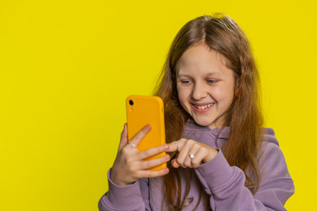 Smiling preteen child girl kid using mobile smartphone typing new post on web, sms message watching video, browsing, addiction of social media networks. Teenager children isolated on yellow backgroundの写真素材