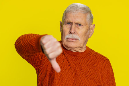 Dislike. Upset unhappy senior old man showing thumbs down sign gesture, expressing discontent, disapproval, dissatisfied negative feedback. Mature pensioner grandfather isolated on yellow backgroundの写真素材