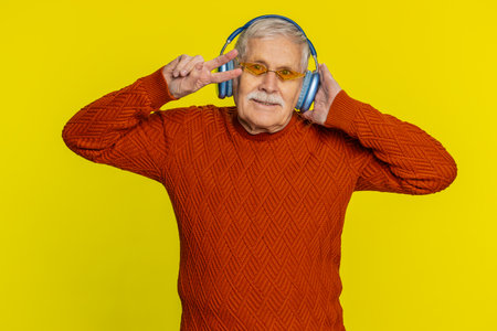 Excited senior stylish old man listening music via headphones and dancing disco party fooling around having fun expressive gesticulating hands. Happy mature grandfather pensioner on yellow backgroundの写真素材