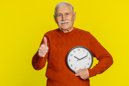 It is your time, hurry up. Senior old man showing time on wall office clock, ok, thumb up approve pointing finger at camera, advertisement, deadline. Elderly grandfather isolated on yellow backgroundの写真素材