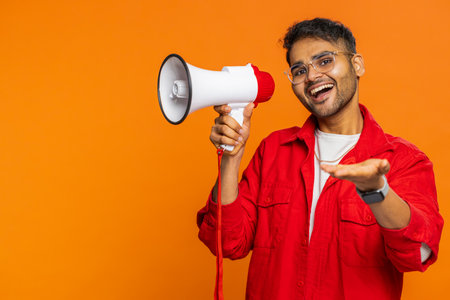 Indian young man talking with megaphone, proclaiming news, loudly announcing advertisement discounts sale using loudspeaker to shout speech. Happy Arabian guy isolated on orange background. Copy-spaceの写真素材