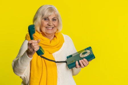 Hey you, call me back. Senior old woman talking on wired landline vintage telephone of 80s, advertising proposition of conversation, online shopping, hotline. Mature grandmother on yellow backgroundの写真素材