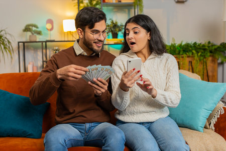 Happy Indian couple holding money cash and smartphone planning discussing investment together...