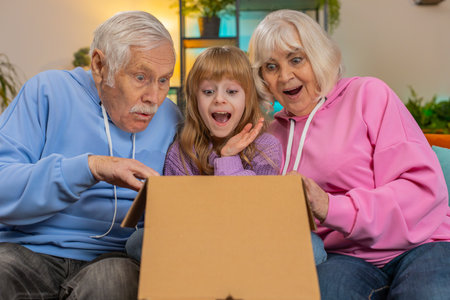 Happy excited Caucasian grandparents and granddaughter girl opening cardboard box together sits on sofa at home. Shocked family consumers unpack good parcel looking inside great purchase delivered.の写真素材