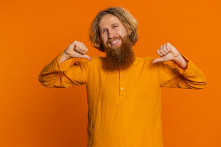 I am the best, choose me. Happy Caucasian man feeling very proud pointing himself, looking self-confident, overjoyed by success, making choice. Young guy isolated on studio orange background indoorsの写真素材