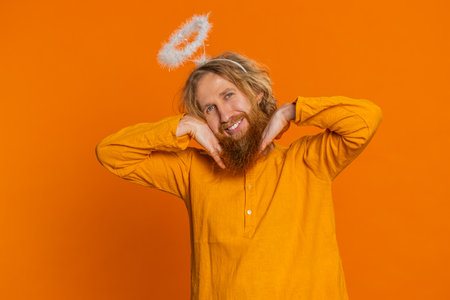 Portrait of smiling shy angelic Caucasian man with angel halo nimb over head flirting, looking at camera, positive emotions, celebrating holidays. Redhead young guy isolated on orange backgroundの写真素材