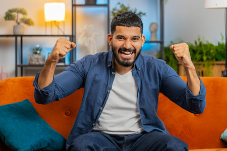 Happy Indian young man watching TV shouting, celebrating success, winning, goal achievement good...