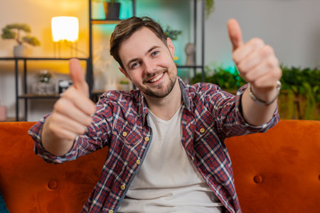 Portrait of excited Caucasian man looking approvingly at camera showing double thumbs up, like sign satisfied, good news, positive feedback. Young happy guy sitting on couch in living room at homeの写真素材