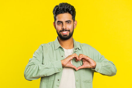 Man in love. Smiling attractive Indian man makes heart gesture demonstrates love sign expresses good positive feelings and sympathy. Handsome Arabian Hindu young guy isolated on yellow wall backgroundの写真素材