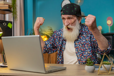 Happy senior old man winner in trendy shirt and cap at home office with laptop scream in triumph gesture celebrate success win money in lottery. Excited elderly mature grandfather get online good newsの写真素材