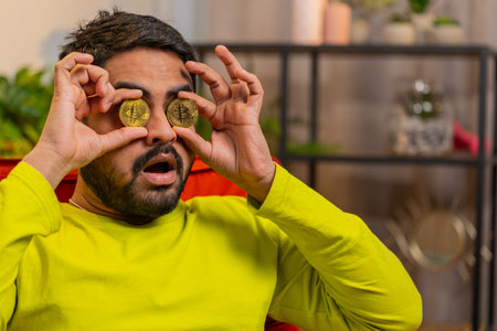Indian man freelancer holding two gold coins, successful developer programmer. Guy stock trader earning bitcoins after online monitoring trading operations. Increasing wealth, financial prosperityの写真素材