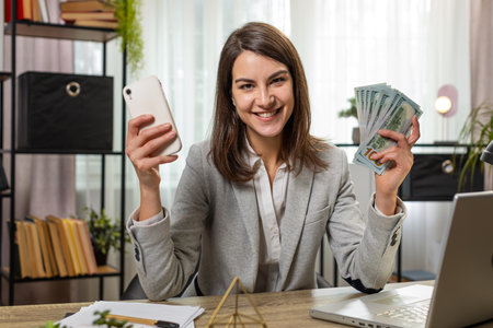 Happy rich businesswoman counting money cash, using smartphone calculator in home office. Successful smiling girl getting bundle of money in office calculates dividends cash money profits financesの写真素材