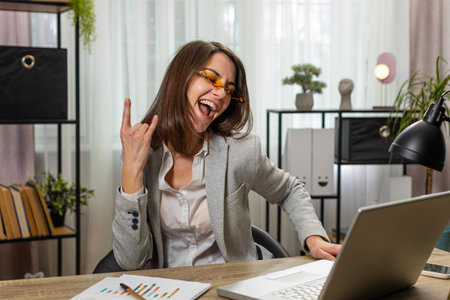 Happy excited Caucasian businesswoman working on laptop at home office desk putting on earbuds listening to favorite energetic disco music and dancing. Freelancer girl relaxing, taking a break.の写真素材