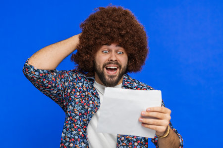 Man with Afro hairstyle wig open envelope take out letter reads it feel happy on blue background. Career growth advance promotion bank loan approve monetary award long-awaited invitation great newsの写真素材