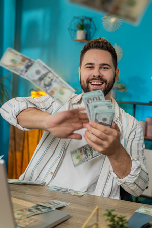Cheerful rich Caucasian young freelancer man working on laptop and wasting throwing money dollar cash more tips earnings big profit success win lottery shopping. Guy sitting at home office. Verticalの写真素材