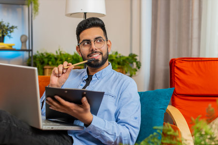 Happy man freelancer work with documents on home sofa reads information business papers prepare financial report on laptop. Indian guy accountant businessman relaxing after finishing work. Copy-spaceの写真素材