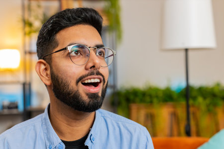 Portrait of amazed shocked Indian man wearing eyeglasses with wow reaction open mouth in living room at home. Happy surprised Arabian guy with big eyes, excited by sudden victory, good win news.の写真素材